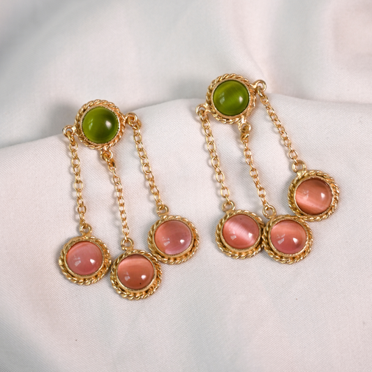 Gold-plated dangle earrings with green and translucent pink stones arranged on white fabric.
