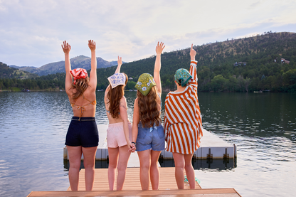 Group wearing colorful bandanas on a lakeside dock, including the Clara bandana in olive green with periwinkle floral details.
