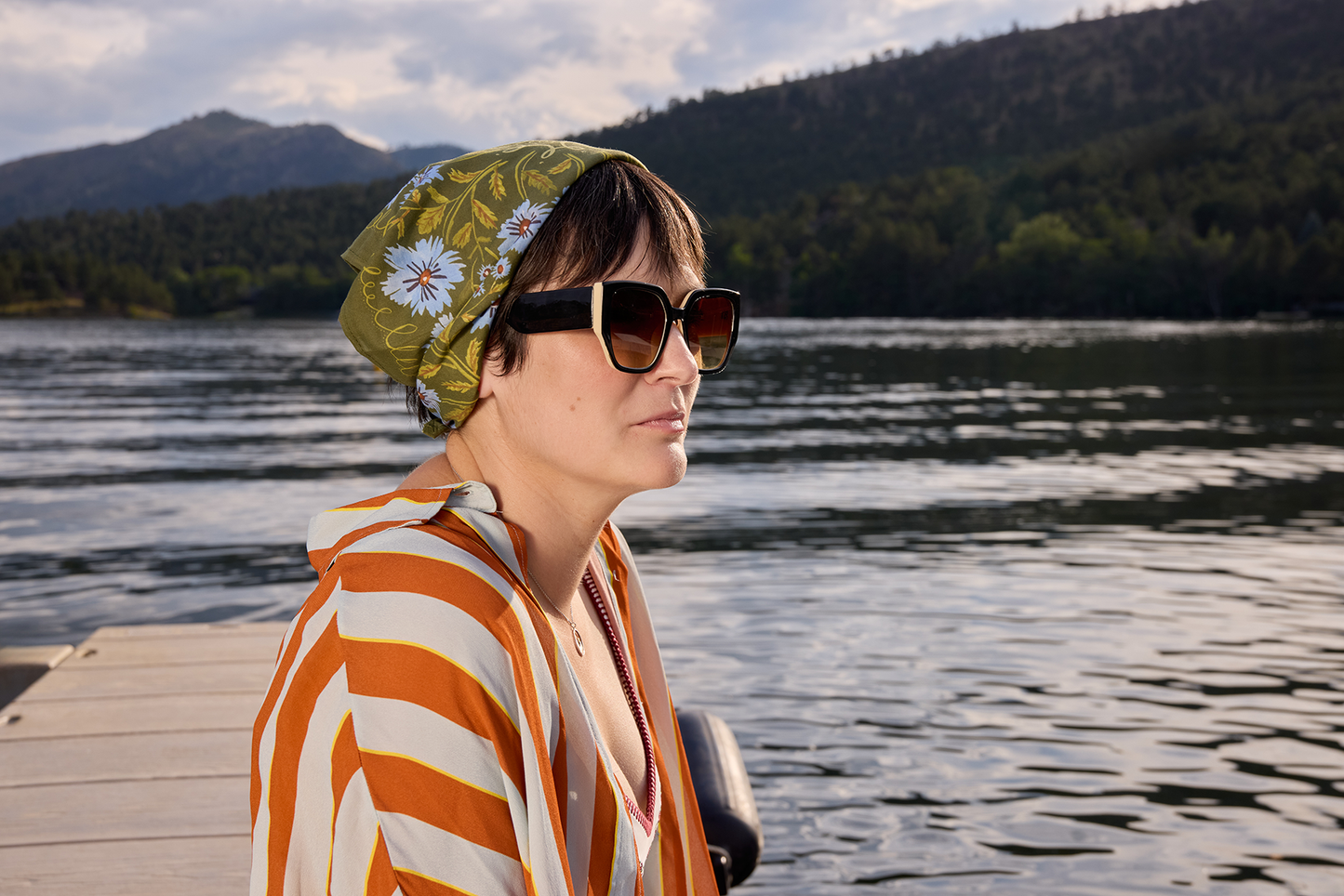 Clara bandana worn as a head scarf by the lake, showing its olive green color and soft periwinkle floral print.
