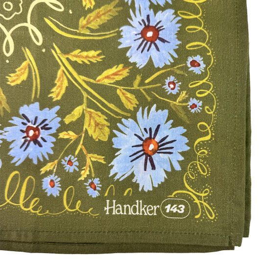 Close-up of the Clara bandana showing periwinkle flowers, olive green cotton fabric, yellow botanical details, and stitched edges.