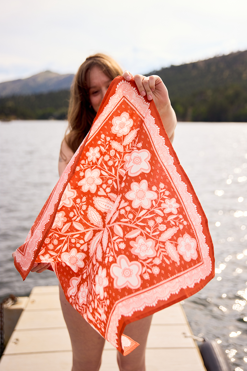 Woman holding the red-orange Lacey Bandana in front of her while standing beside a mountain lake.