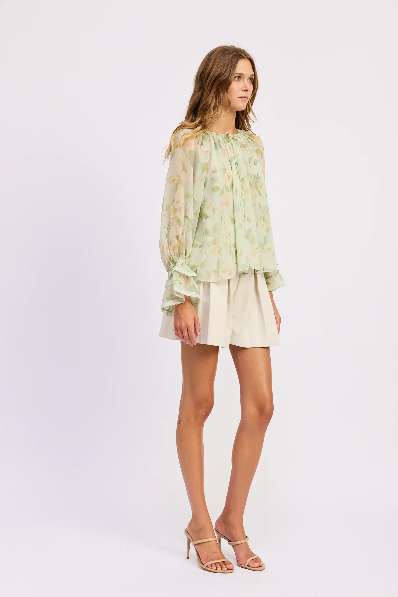 Model wearing the Paloma Blouse in a sheer light green floral print with a gathered tie neckline, long sleeves, and ruffled cuffs styled with cream shorts.