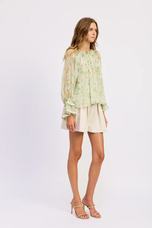 Model wearing the Paloma Blouse in a sheer light green floral print with a gathered tie neckline, long sleeves, and ruffled cuffs styled with cream shorts.