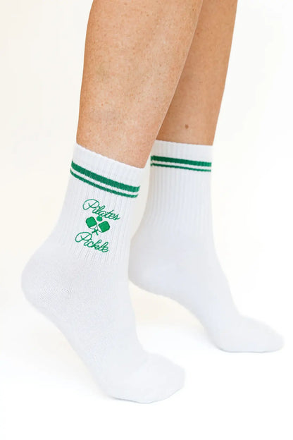 Close-up of white Pilates Pickle crew socks featuring green varsity stripes and an embroidered pickleball graphic with script lettering.