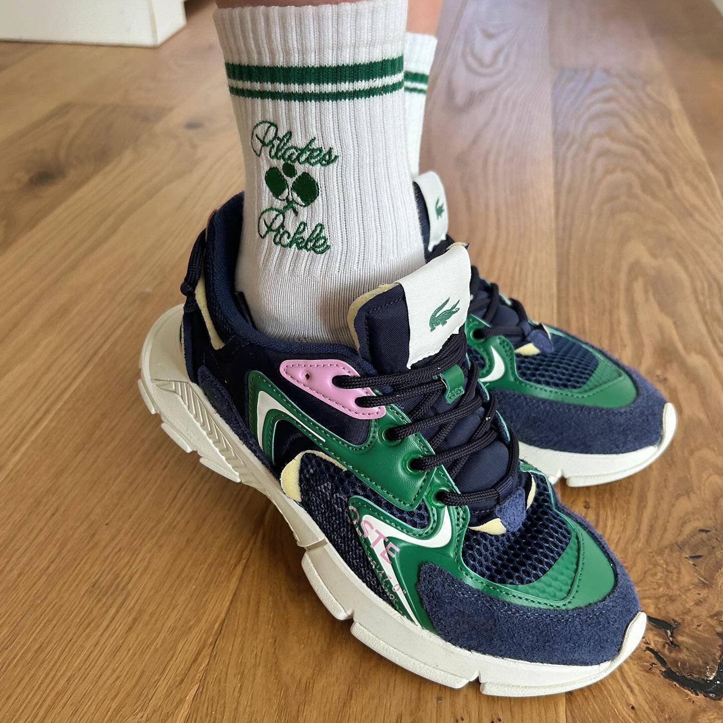 Close-up of white Pilates Pickle socks featuring green embroidered lettering, pickleball motif, varsity stripes, and colorful athletic sneakers.