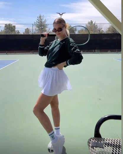 Woman standing on a tennis court wearing a pleated white skirt, sweatshirt, and white Pilates Pickle socks with green stripe accents.