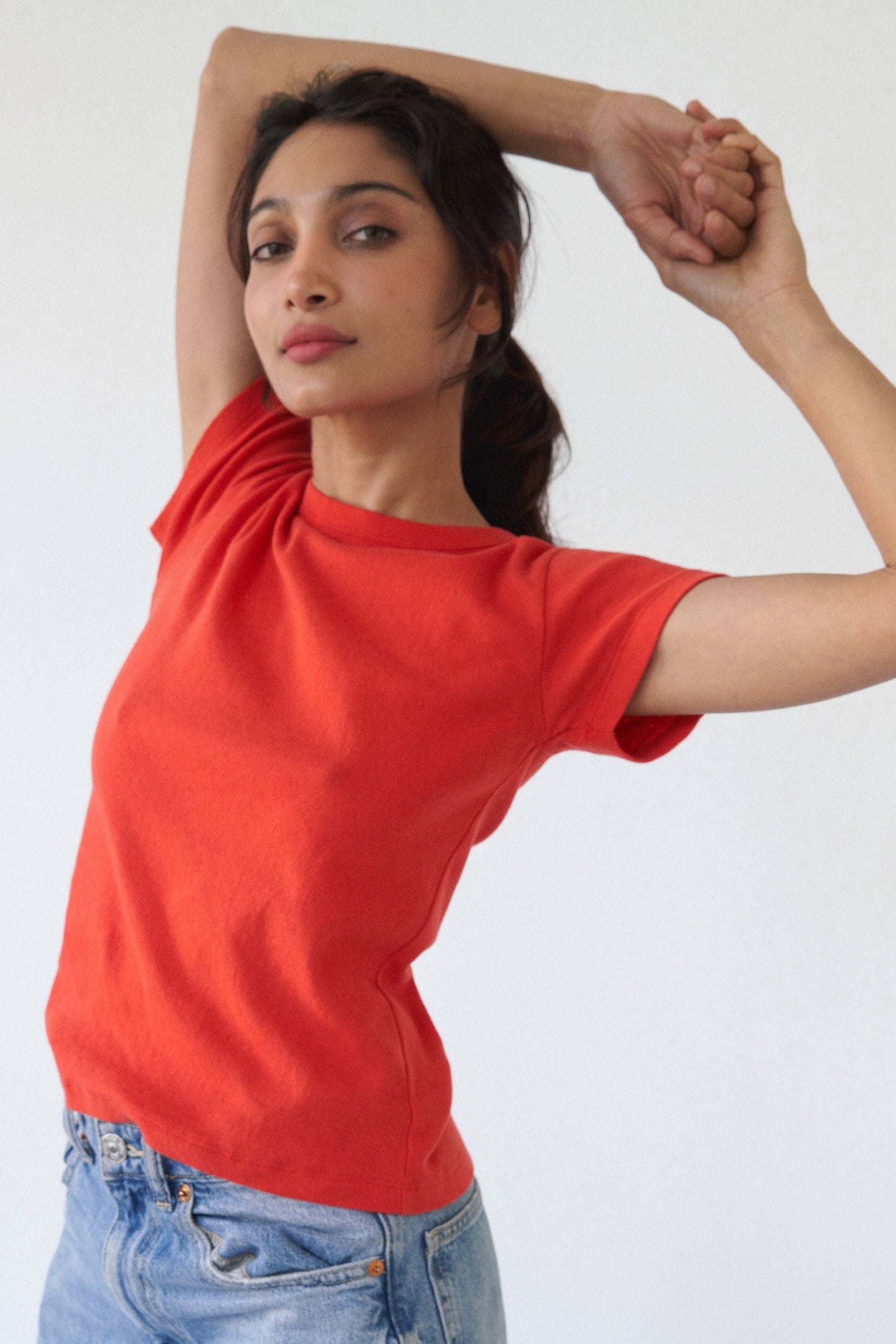 Model wearing the Pointelle Baby Tee in red orange with light wash jeans, shown from the front with arms raised.