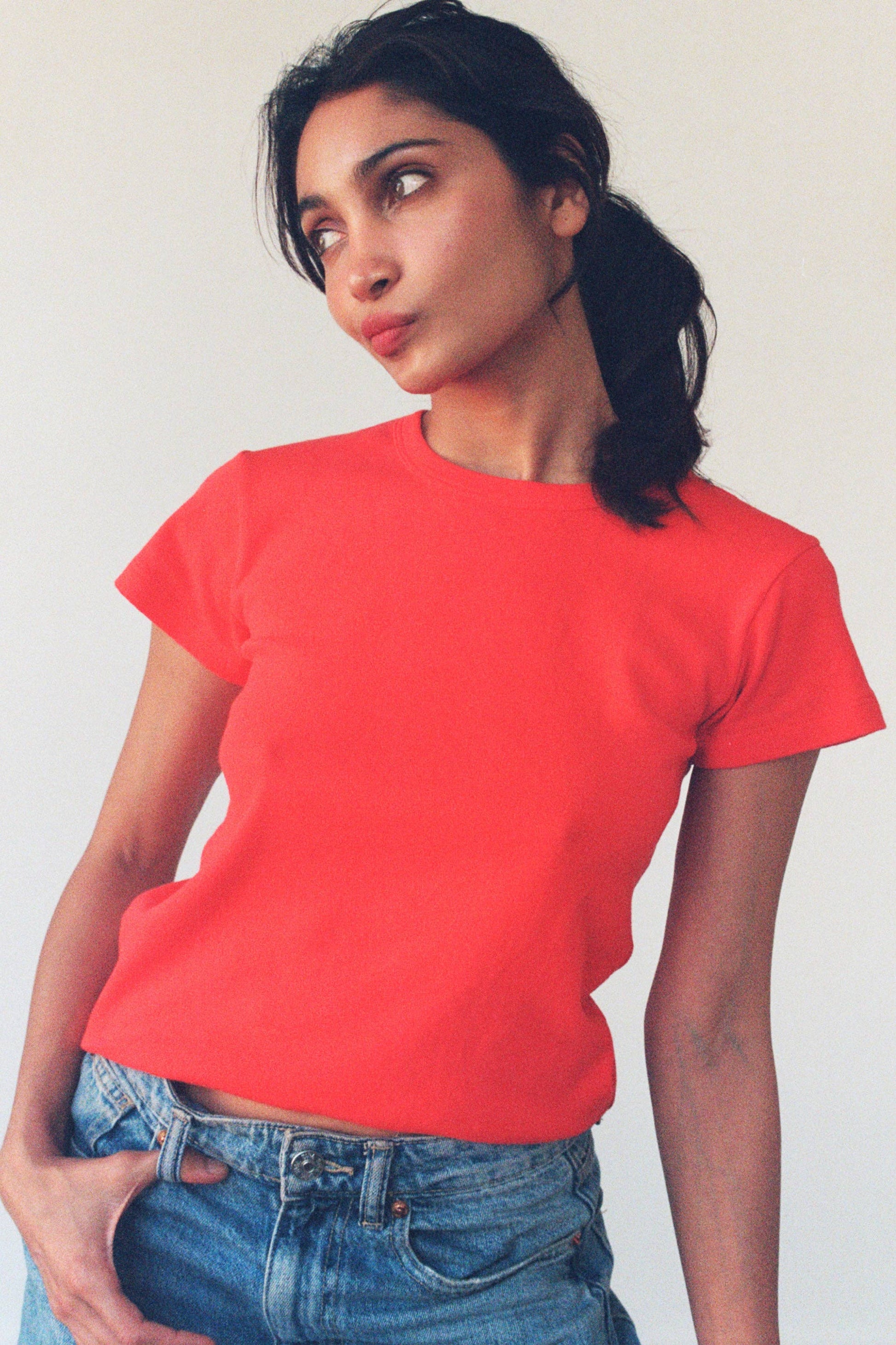 Model styling the Pointelle Baby Tee in red orange tucked into light wash jeans, showing the slightly shrunken fit.