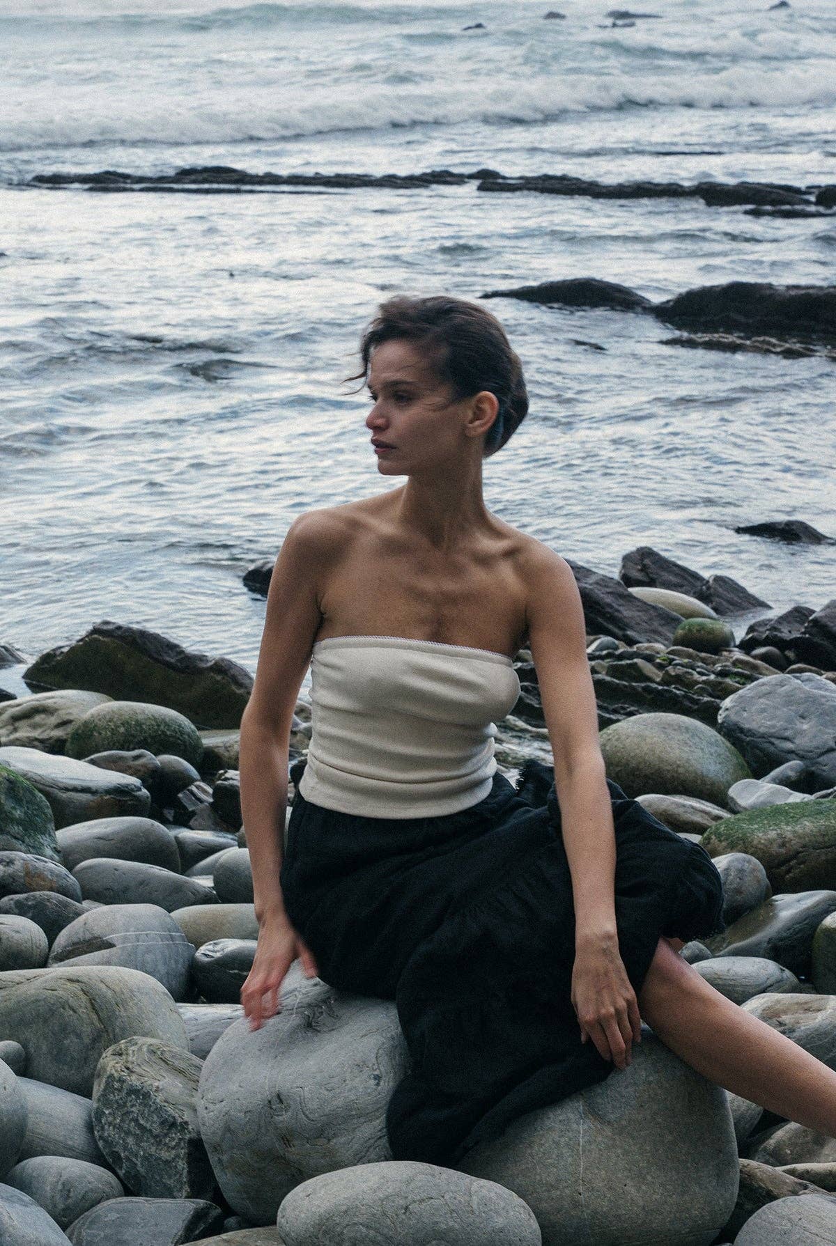 Model wearing the Pointelle Tube Top in Creme with a black flowing skirt while seated on coastal rocks by the ocean.