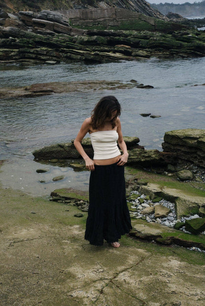 Full-length view of the Pointelle Tube Top in Creme paired with a flowing black skirt beside a rocky shoreline.