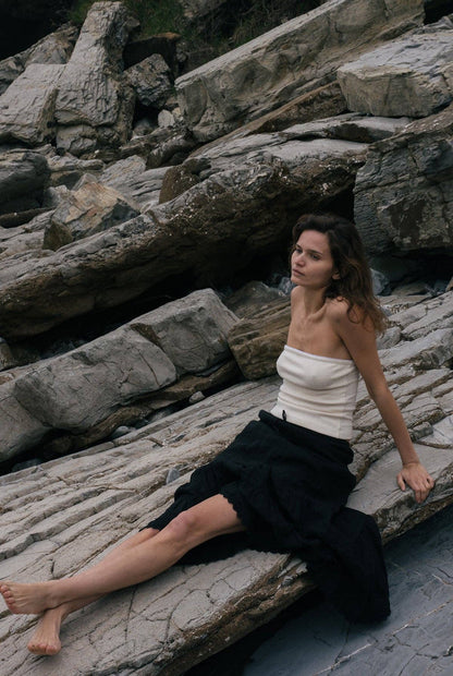 Model wearing the creme pointelle tube top styled with a black skirt while seated against layered coastal rocks.