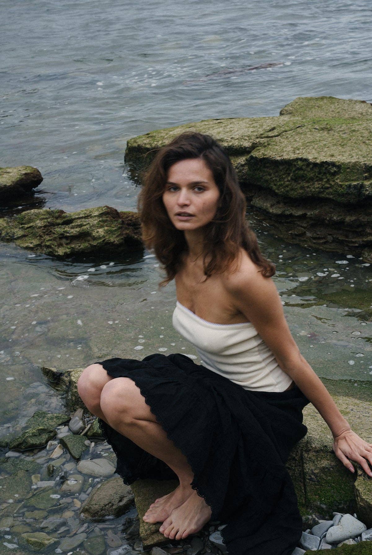 Model crouching near the water wearing the creme pointelle tube top and a black textured skirt.