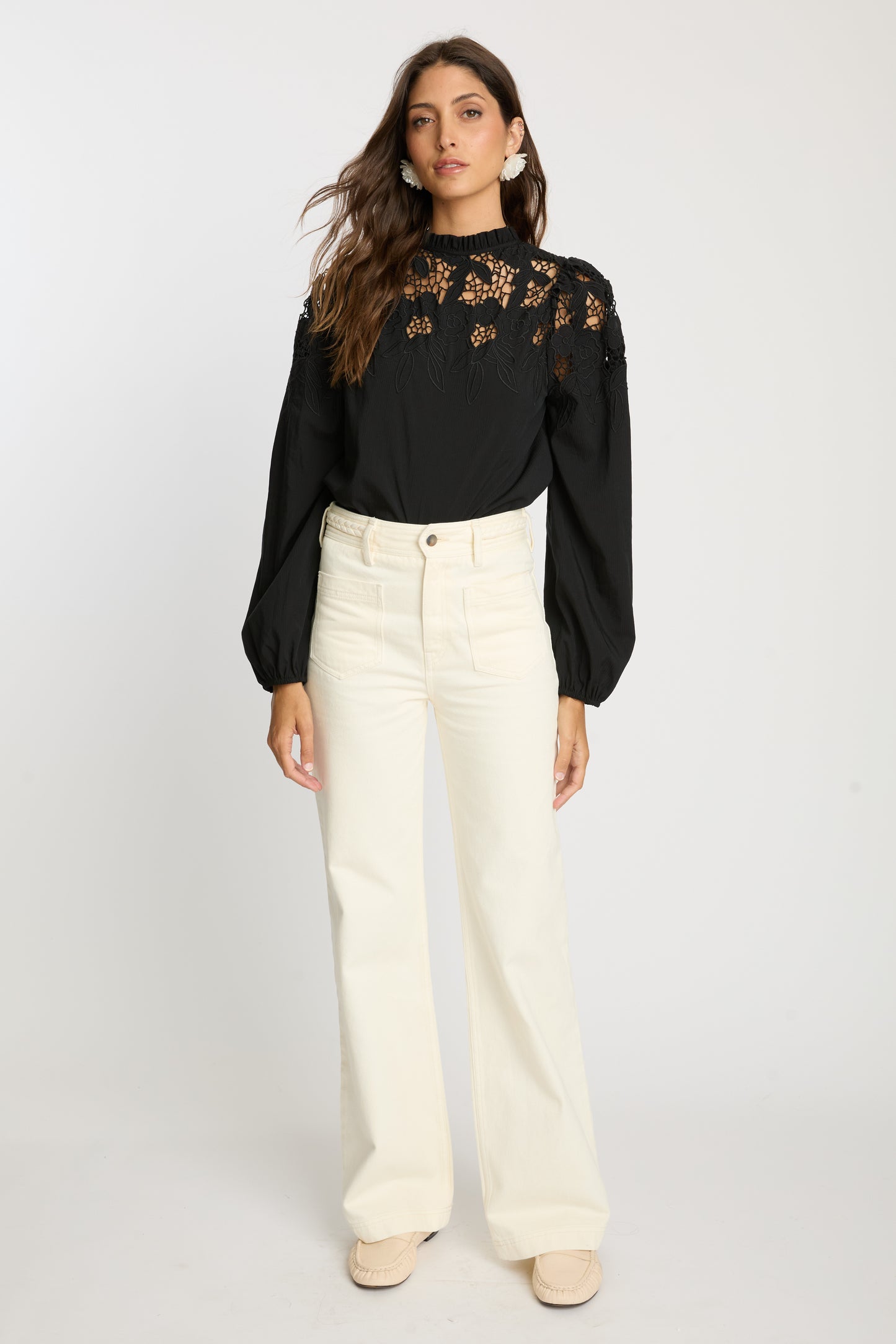 Model wearing the Riana Reversible Top in black with cream wide-leg pants, featuring a high ruffled neckline, sheer embroidered lace bodice, and voluminous long sleeves.