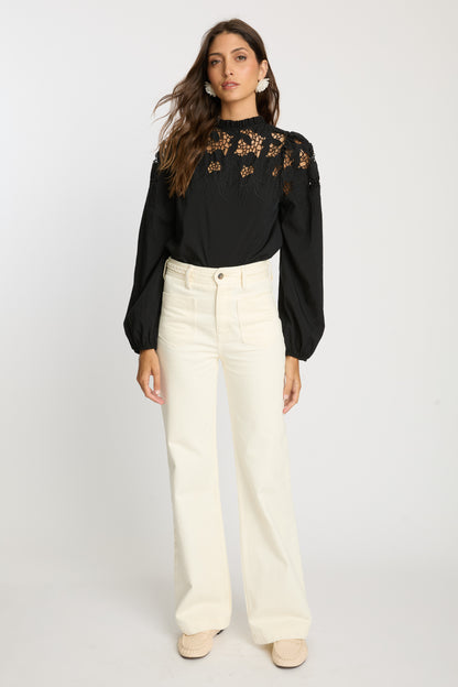 Model wearing the Riana Reversible Top in black with cream wide-leg pants, featuring a high ruffled neckline, sheer embroidered lace bodice, and voluminous long sleeves.