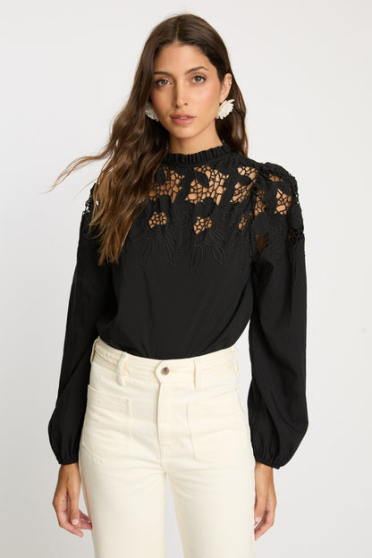 Model wearing the Riana Reversible Top in black featuring intricate floral lace embroidery, a high ruffled neckline, semi-sheer upper bodice, and long gathered sleeves.