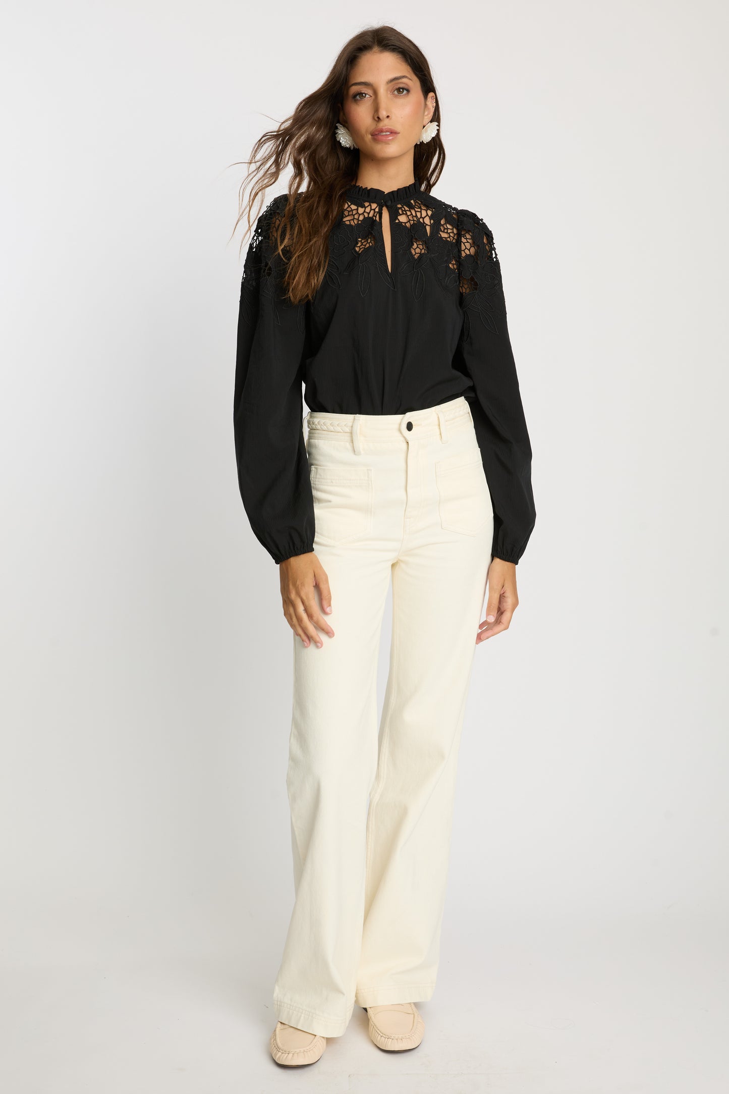Model wearing the Riana Reversible Top in black styled with cream wide-leg pants, featuring floral lace embroidery, a ruffled neckline, sheer upper panels, and long gathered sleeves.