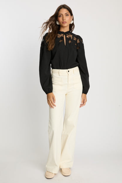 Model wearing the Riana Reversible Top in black styled with cream wide-leg pants, featuring floral lace embroidery, a ruffled neckline, sheer upper panels, and long gathered sleeves.