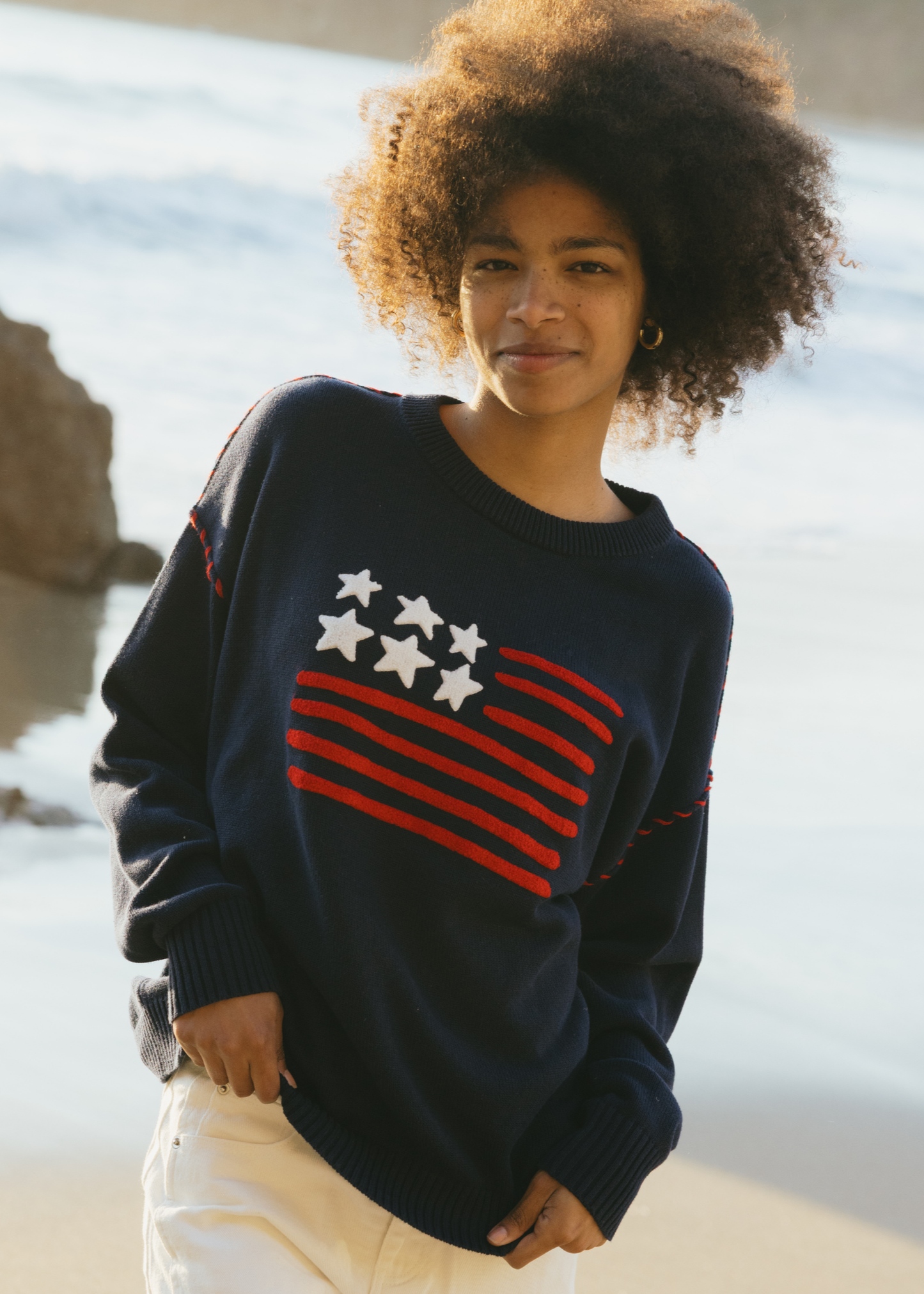 Model wearing the navy Riley Crew sweater with an American flag design, styled with light pants outdoors.