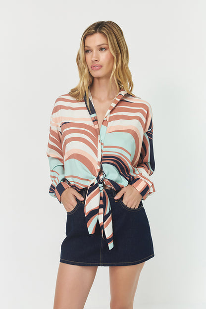 Model wearing the Robbie Top in Wavy Geo print featuring pink, blue, and earthy swirl patterns, satin fabric, long sleeves, and a front tie paired with a dark denim mini skirt.