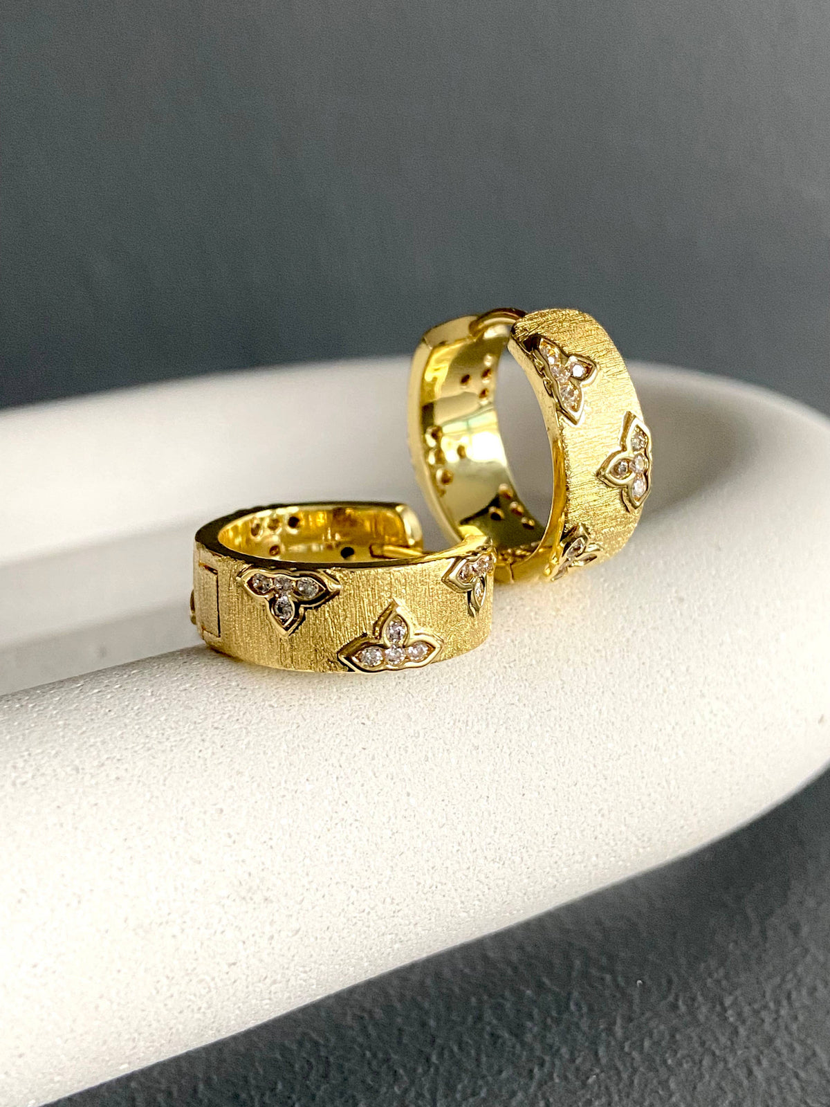Satia gold hoop earrings with matte ribbed texture and rhinestone four-leaf clover details displayed on a jewelry cushion.