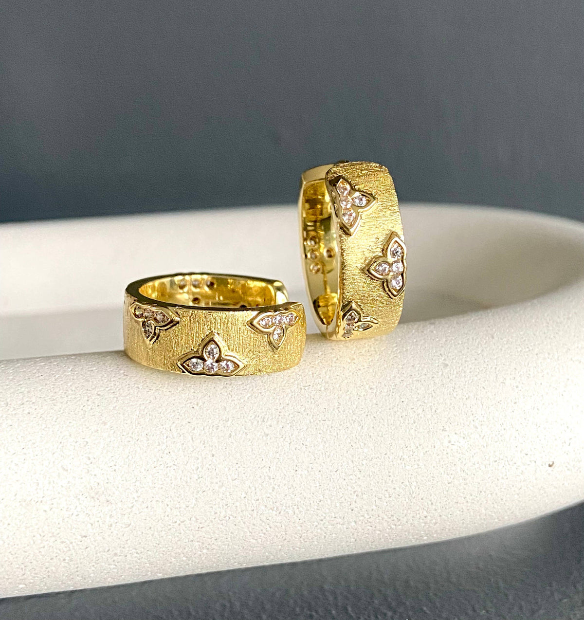 Pair of Satia 18K gold-plated hoop earrings with ribbed finish and rhinestone clover accents displayed on a cushion.