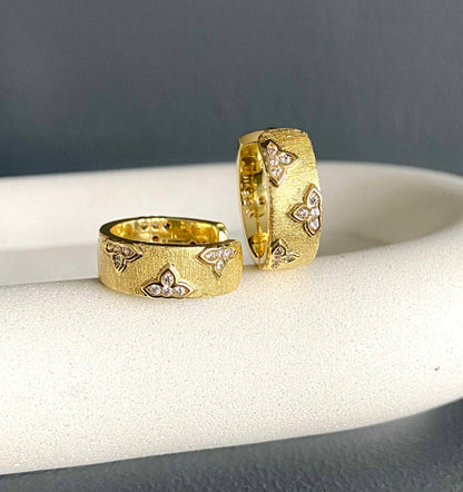 Pair of Satia 18K gold-plated hoop earrings with ribbed finish and rhinestone clover accents displayed on a cushion.
