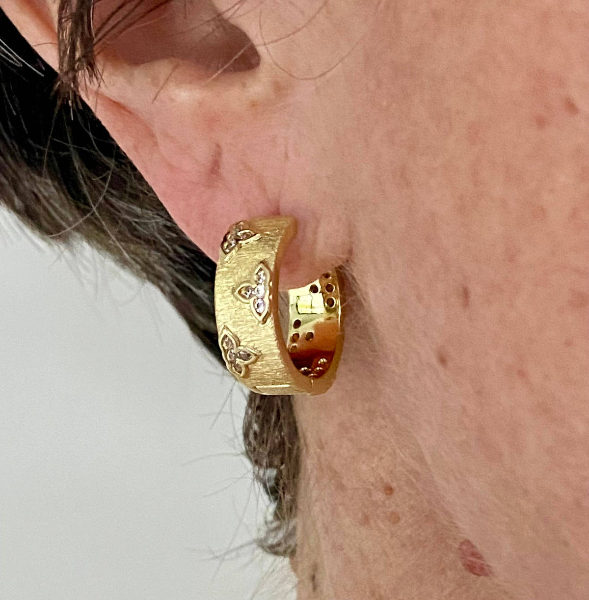 Satia gold hoop earring worn on ear, featuring a matte textured finish with delicate rhinestone clover accents.