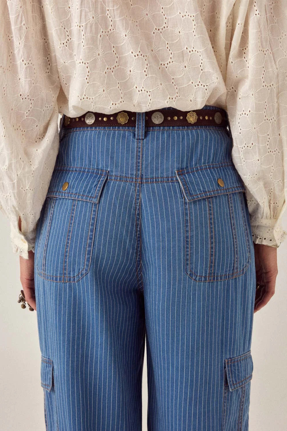 Model wearing the Sikka Belt in Tobacco with blue pinstripe denim trousers and a cream embroidered blouse, showcasing the coin-detailed suede leather belt.
