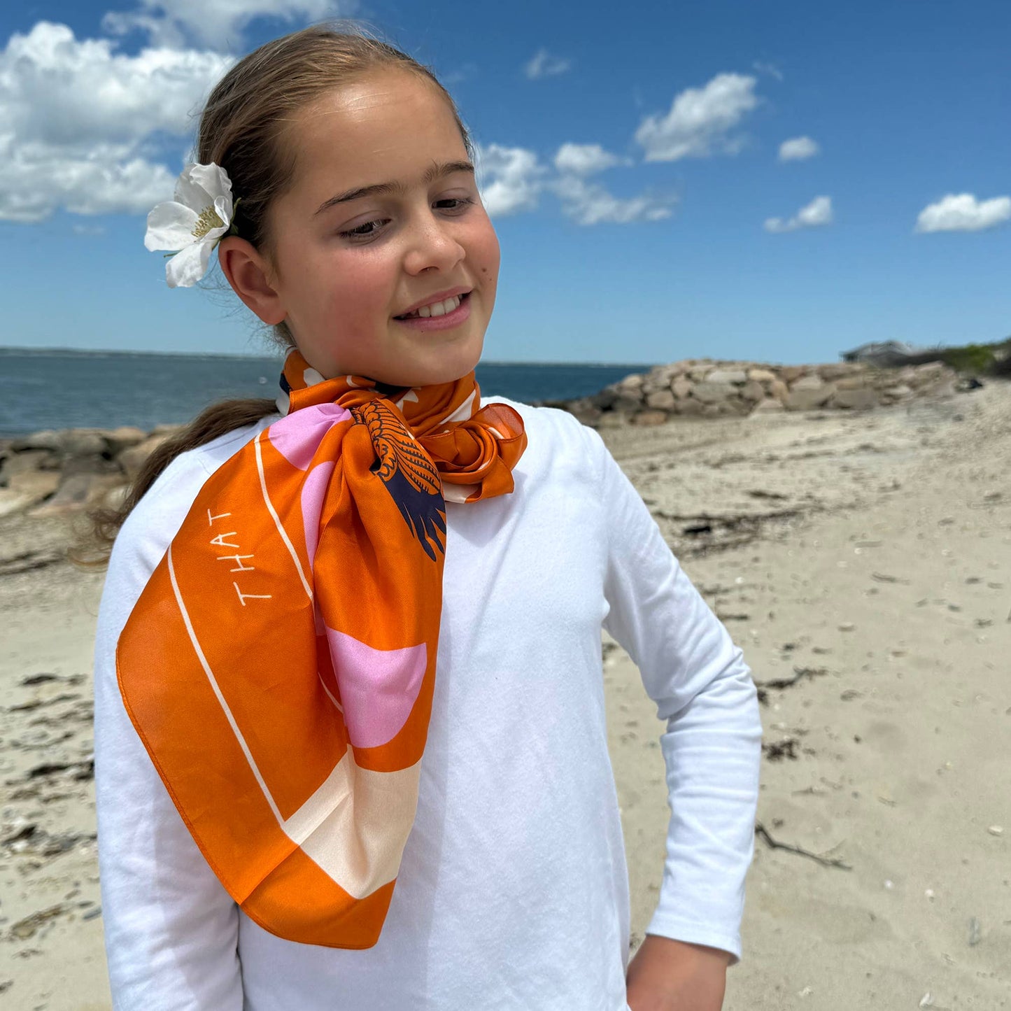 Child model at the beach wearing the Soar & Sing silk scarf tied around the neck with a white long sleeve top.