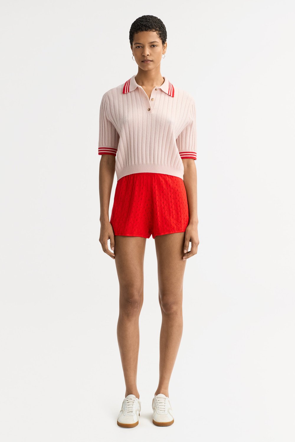 Full-length front view of the blush pink Striped Pique cashmere polo styled with bright red knit shorts and white sneakers.