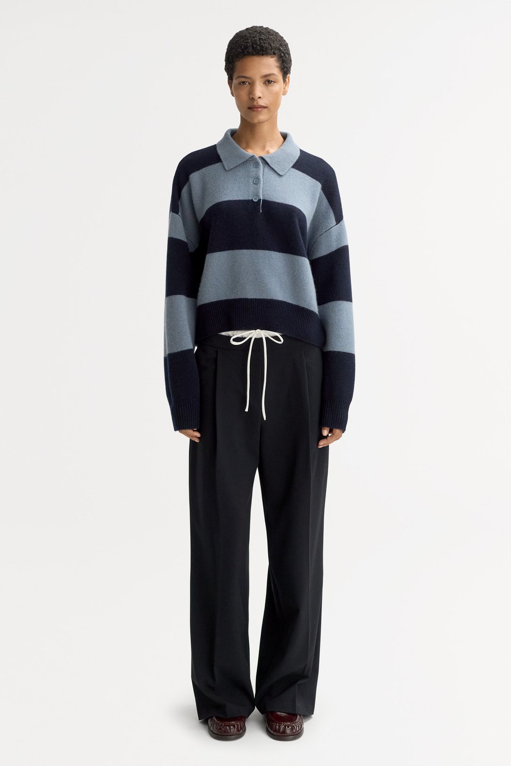Model wearing the Striped Polo Shirt in Retro Blue with wide-leg black trousers, showcasing the cropped silhouette, polo collar, and chunky striped cashmere knit.
