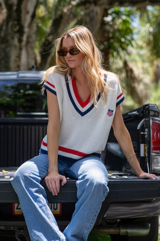 Model seated outdoors wearing the American Shield sweater vest with light wash jeans.