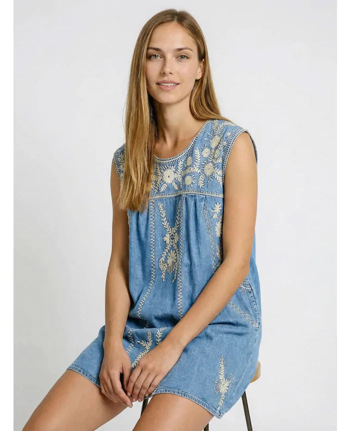 Seated model wearing the Sylvie Arizona embroidered denim mini dress with white embroidered detailing and sleeveless design