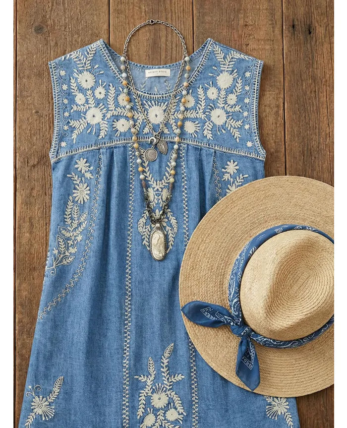 Sylvie Arizona embroidered denim mini dress styled with layered necklaces and straw hat on wooden background