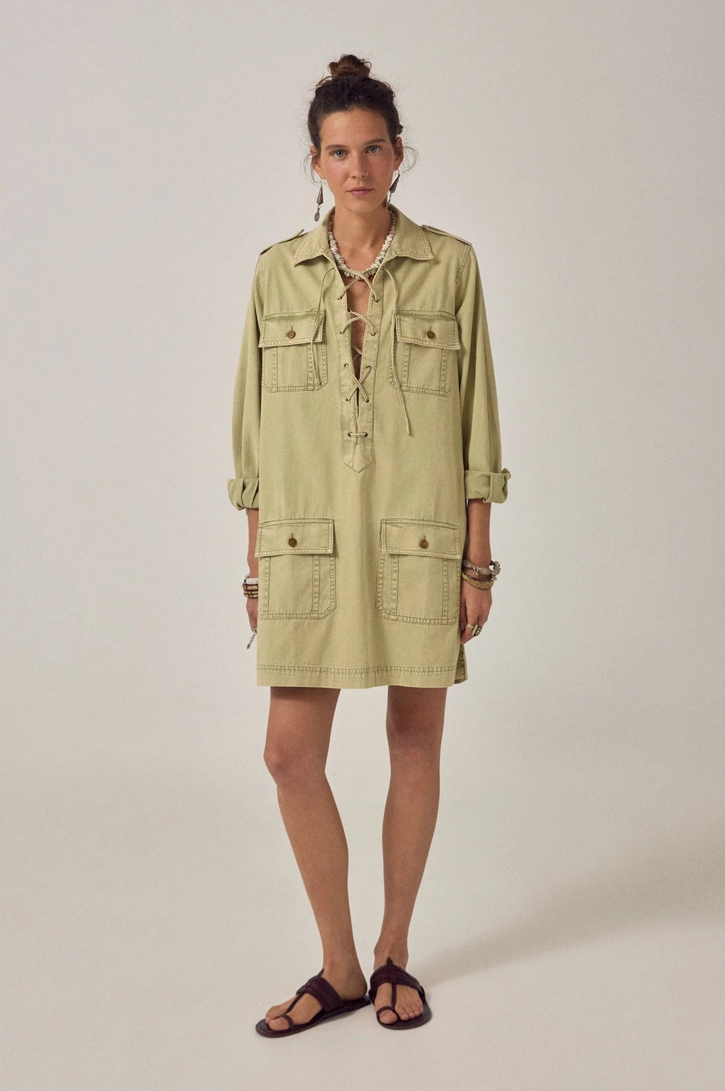Model wearing the Thalassa Short Dress in Army Khaki featuring a lace-up V-neckline, long sleeves with epaulets, and four flap pockets.