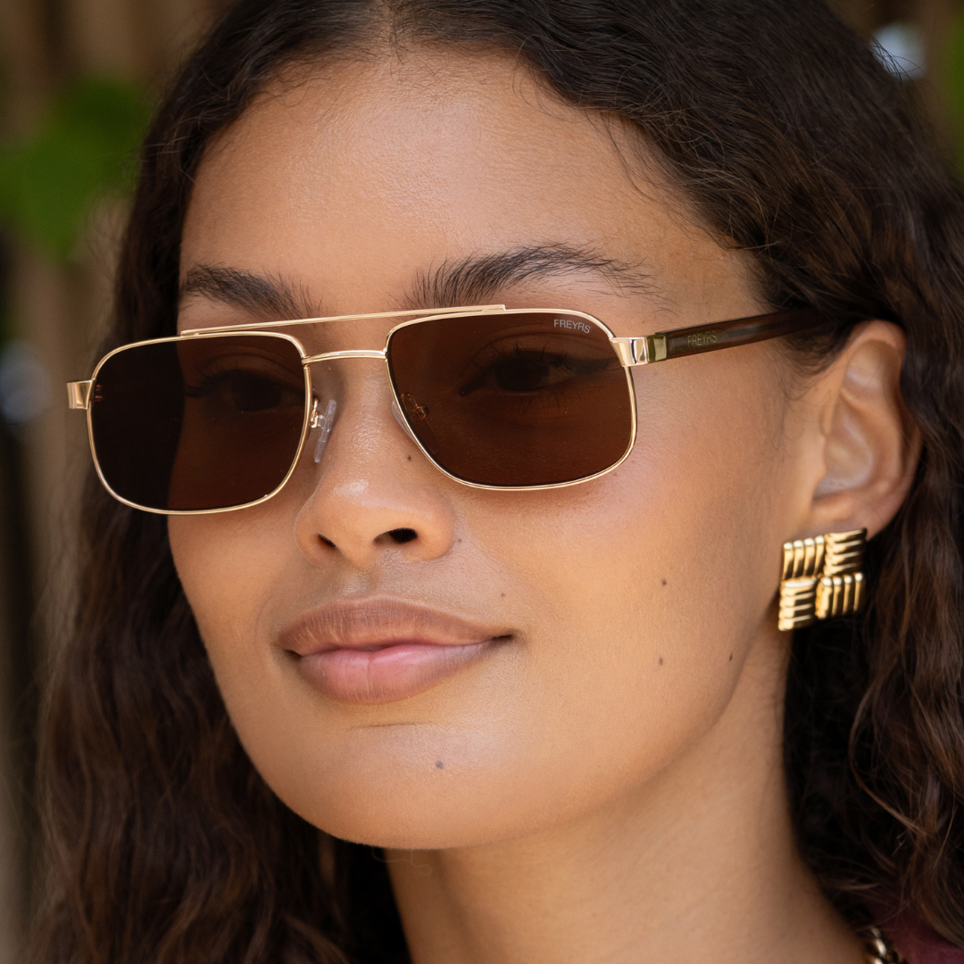 Model wearing Vesper sunglasses with a gold-tone metal frame, brown lenses, and tortoise temples.