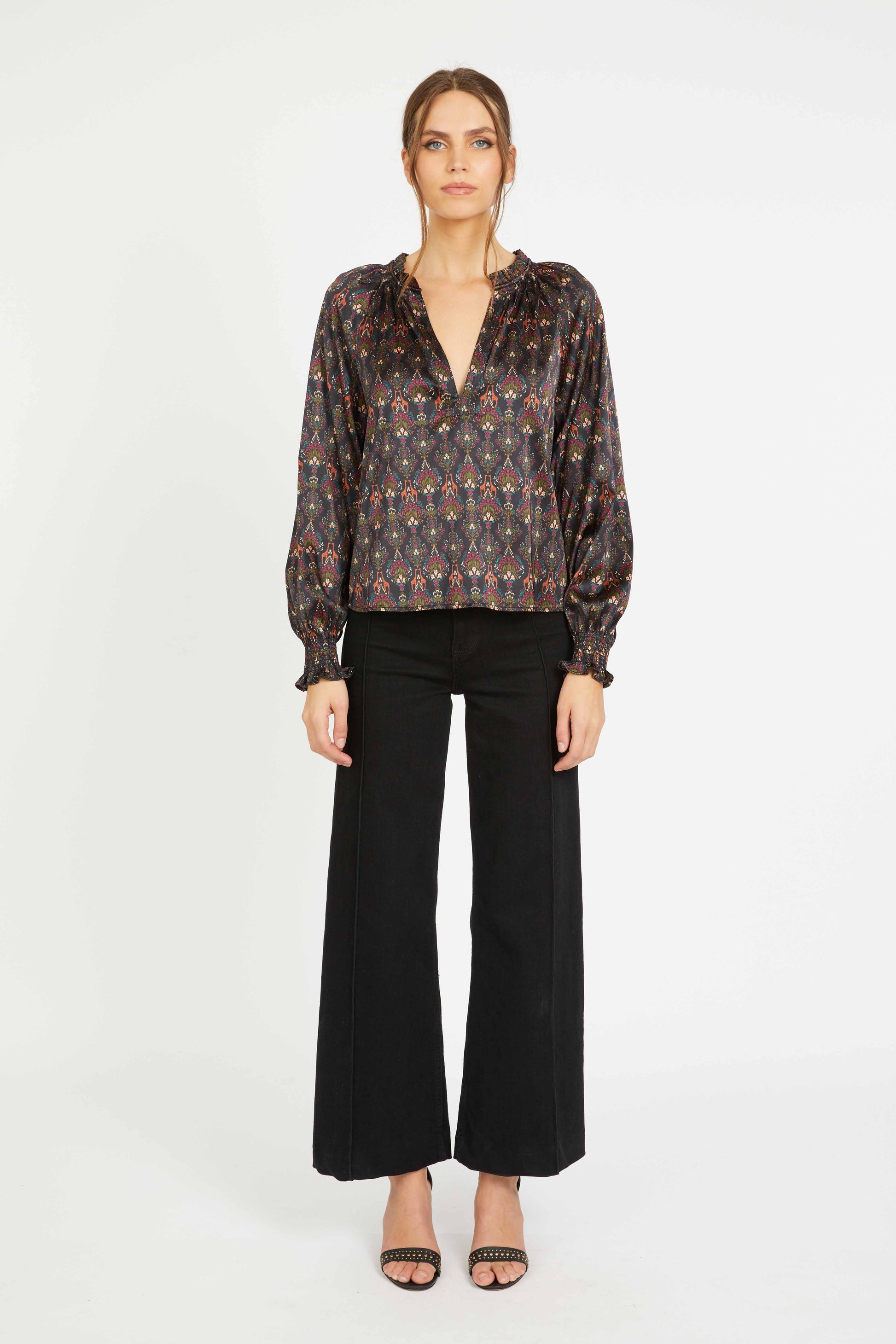 Model wearing the Vickie Top in a dark medallion-inspired satin print with a V-neckline, gathered collar, long voluminous sleeves, and ruffled cuffs paired with black wide-leg pants.