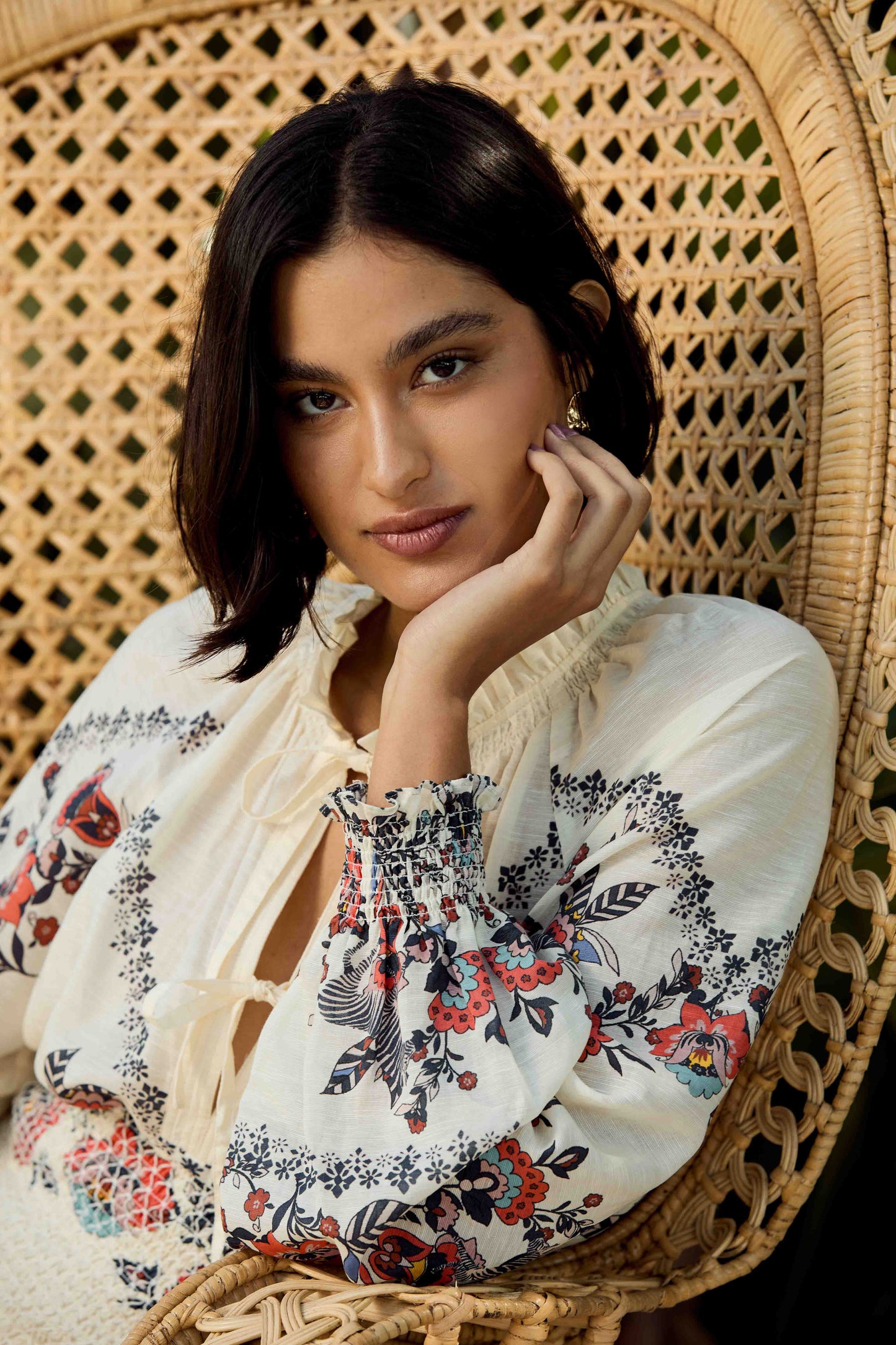 Portrait of model seated in a wicker chair wearing the Vickie Top in Zebra Leaf with floral embroidery, smocked cuffs, and tassel tie neckline in a warm resort-inspired setting.