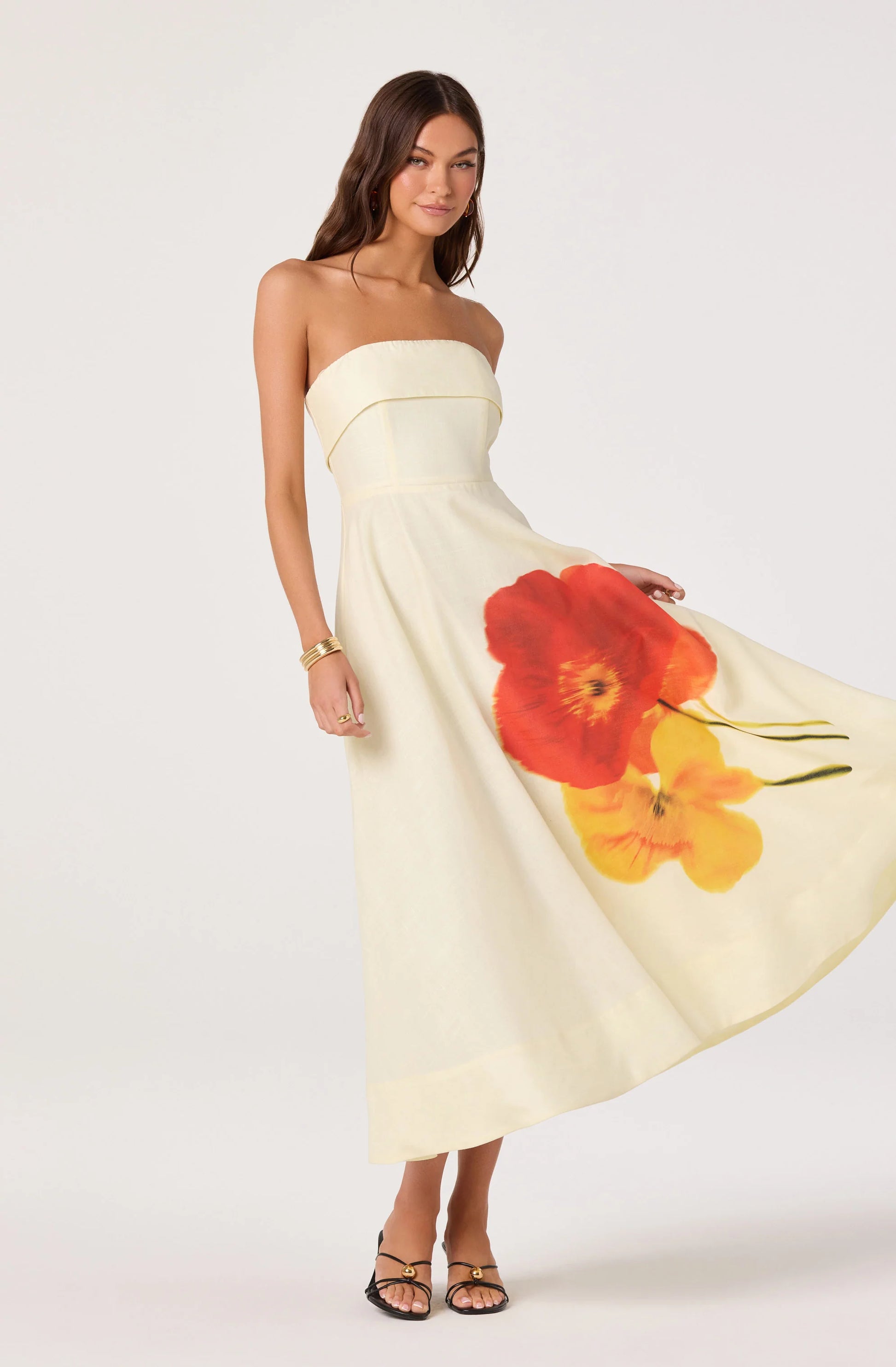 Model holding out the skirt of the Ximena Floral Strapless Midi Dress, showing the A-line shape and large red and yellow floral placement print.